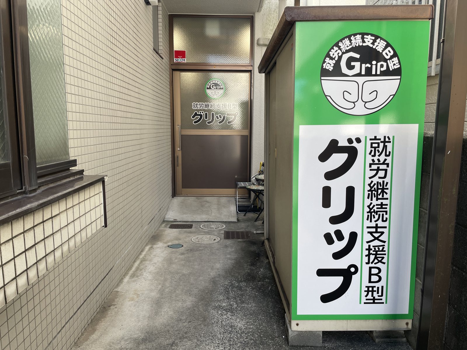 建物の右側の通路の奥にあるグリップの入口と、「就労継続支援B型 グリップ」の看板